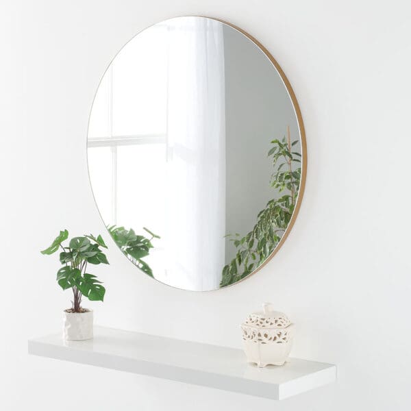 Frameless all glass round mirror. Features a gold backing. Wall mounted above a shelf. Reflecting a window.