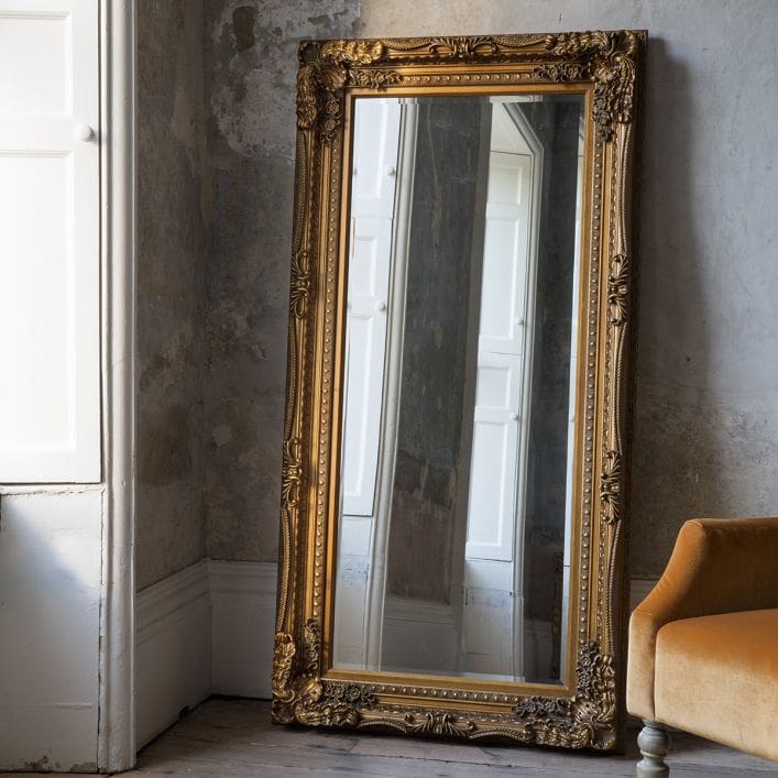 Large full length framed mirror. Features a gold decorative frame, leaning against a living room wall reflecting the interior.
